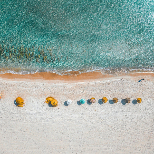 Drone photo of an empty beach in Miami Beach, FL. The water is calm and turquoise colored, with various umbrellas placed on the light yellow sand